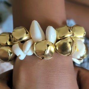 May include: A gold-toned bracelet featuring alternating gold bells and white seashells. The bracelet is worn on a wrist, showcasing the contrasting textures and colors of the jewelry. The bells have a small slit, and the shells are smooth.