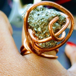 May include: A copper wire ring with a large, irregularly shaped pyrite stone wrapped in the wire. The ring is on a person's finger.
