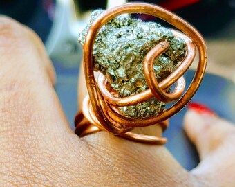 May include: A copper wire ring with a large, irregularly shaped pyrite stone wrapped in the wire. The ring is on a person's finger.