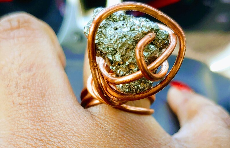 May include: A copper wire ring with a large, irregularly shaped pyrite stone wrapped in the wire. The ring is on a person's finger.