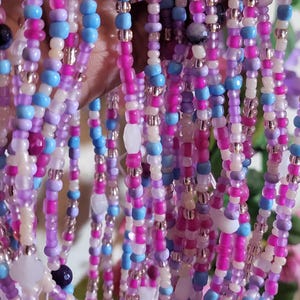 May include: Strands of colorful beads in shades of pink, purple, blue, and white. The beads are strung together, creating a textured and layered effect. Some beads have a metallic sheen. The image suggests jewelry or craft supplies.