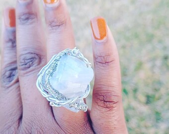May include: A silver wire wrapped ring featuring a large, white, opaque, natural crystal. The ring is worn on a person's finger.