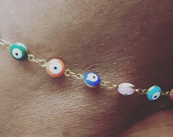 May include: Gold chain anklet with colorful evil eye charms. The charms are round and have a white center with a black pupil. The anklet is made of a thin gold chain and has a lobster clasp closure.