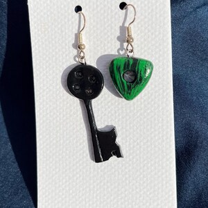 May include: A pair of handmade earrings. One earring is a black key shape, and the other is a green and black triangle. Both earrings have silver-colored ear wires. The earrings are displayed on a white card.