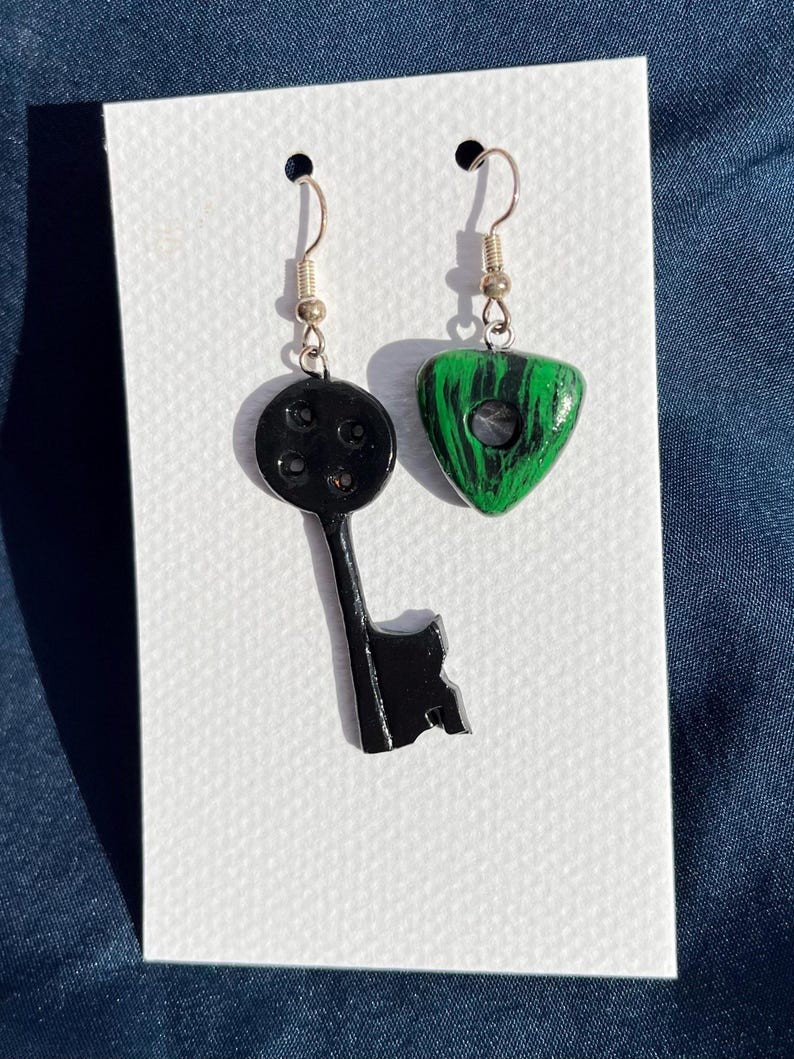 May include: A pair of handmade earrings. One earring is a black key shape, and the other is a green and black triangle. Both earrings have silver-colored ear wires. The earrings are displayed on a white card.