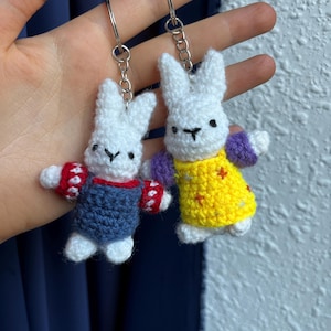 Könnte beinhalten: Zwei kleine, weiße, gehäkelte Hasen-Schlüsselanhänger mit Metallringen. Ein Hase trägt eine blaue Latzhose mit rot-weiß gemusterten Ärmeln. Der andere Hase trägt ein gelbes Kleid mit violetten Ärmeln und kleinen orangefarbenen Sternen.