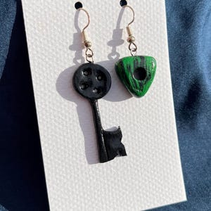 May include: A pair of handmade earrings. One earring is a black key shape with a round top and the other is a green triangle shape with black streaks. Both earrings have silver-colored hooks.