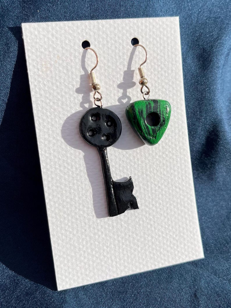 May include: A pair of handmade earrings. One earring is a black key shape with a round top and the other is a green triangle shape with black streaks. Both earrings have silver-colored hooks.