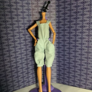 May include: A headless doll wearing a light blue overall jumpsuit with a stand. The doll is standing on a purple stand with three legs.