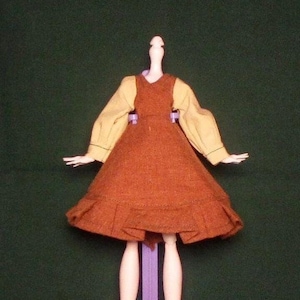 May include: A doll wearing a brown dress with a yellow blouse and a purple stand.