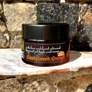 Może przedstawiać: Ciemnobursztynowy szklany słoik z czarną zakrętką. Etykieta zawiera tekst Australian sandalwood cedarwood bergamot patchouli sweet orange oraz Sandalwood Orange z małą grafiką plasterka pomarańczy na czarnym tle.