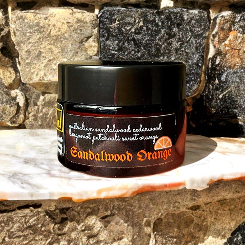 Może przedstawiać: Ciemnobursztynowy szklany słoik z czarną zakrętką. Etykieta zawiera tekst Australian sandalwood cedarwood bergamot patchouli sweet orange oraz Sandalwood Orange z małą grafiką plasterka pomarańczy na czarnym tle.