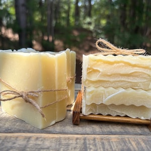 May include: Two rectangular bars of pale yellow soap with wavy, textured tops. One bar stands upright, wrapped in thin brown twine tied in a bow. The other bar rests on a small, dark brown wooden soap dish, also secured with brown twine.