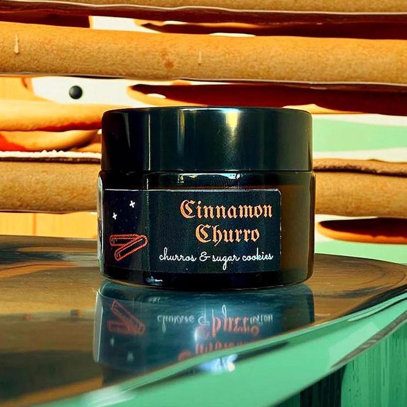 Może przedstawiać: Mały słoik z ciemnego bursztynowego szkła z czarną zakrętką, posiadający czarną etykietę z napisem Cinnamon Churro oraz churros & sugar cookies w pomarańczowej i białej czcionce, wraz z małą grafiką przedstawiającą dwa churros i gwiazdki.