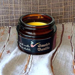 Może przedstawiać: Mały słoiczek z bursztynowego szkła zawierający ż&oacute;łty wosk, z czarną etykietą z napisem "French Vanilla Tobacco Absolute" i "Smoked Vanilla" oraz grafiką przedstawiającą fajkę z czerwoną gł&oacute;wką i białym dymem.