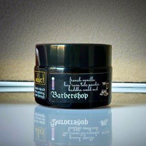 Może przedstawiać: Czarny cylindryczny słoik z etykietą zawierającą tekst Barbershop, french vanilla, bay rum talc powder, buddha wood oud oraz grafikę słupka i krzesła fryzjerskiego. Nazwa marki Addict jest wydrukowana na oddzielnej etykiecie bocznej.