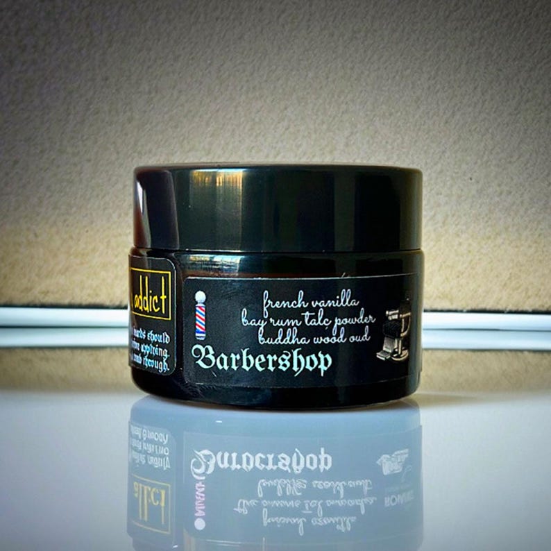 Może przedstawiać: Czarny cylindryczny słoik z etykietą zawierającą tekst Barbershop, french vanilla, bay rum talc powder, buddha wood oud oraz grafikę słupka i krzesła fryzjerskiego. Nazwa marki Addict jest wydrukowana na oddzielnej etykiecie bocznej.