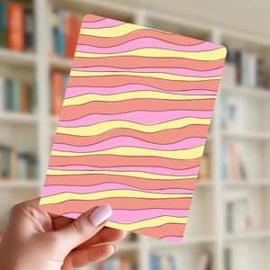 May include: A rectangular card with a wavy pattern in shades of pink, yellow, and peach. The design features horizontal, flowing lines. The card is held in a hand, with a blurred bookshelf in the background.
