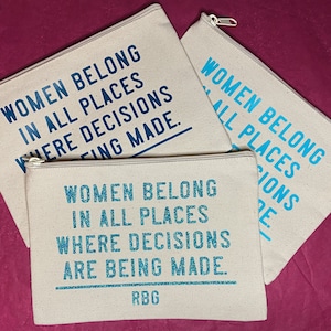 May include: Three beige zippered pouches with the text "Women belong in all places where decisions are being made." The bottom pouch has the initials "RBG" printed below the text.