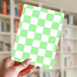 May include: A rectangular card with a green and white checkerboard pattern. The card is held in a hand with a blurred bookshelf in the background. The card's rounded corners and the checkerboard design are clearly visible.