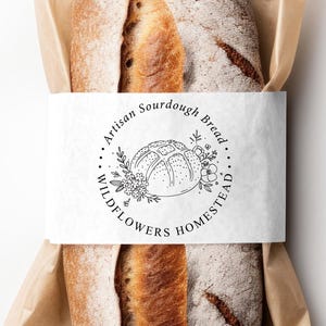 May include: Artisan sourdough bread in a brown paper bag, with a white label wrapped around the loaf. The label reads "Artisan Sourdough Bread Wildflowers Homestead" in a circular design with floral illustrations.
