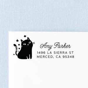 May include: A black ink stamp impression on white paper featuring a stylized black cat with stars and the text Amy Parker, 1496 LA SIERRA ST, MERCED, CA 95348. The cat has a striped tail and simple facial features, positioned to the left of the address.