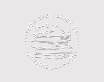 Op de afbeelding: Een cirkelvormig stempelontwerp met een witte lijntekening van een kat die op een stapel van vier boeken slaapt. De tekst luidt FROM THE LIBRARY OF en ADELINE JOHNSON in een cirkelvormige rand met kleine stippen als scheiding.