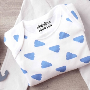 K&ouml;nnte beinhalten: Ein wei&szlig;er kurz&auml;rmeliger Babybody mit einem sich wiederholenden Muster aus hellblauen Wolken. Im Bereich des Halsausschnitts ist der schwarze Text Adeline JOHNSON in einer einfachen Schriftart aufgedruckt. Das Kleidungsst&uuml;ck hat einen klassischen Schlupfausschnitt.