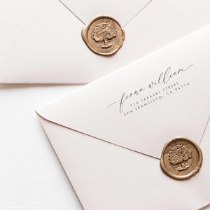 May include: Two light pink envelopes sealed with metallic gold wax seals featuring a tree design. One envelope displays the printed black text: fiona william, 719 TARAVAL STREET, SAN FRANCISCO, CA 94116 in a combination of script and sans-serif fonts.