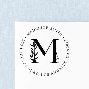 May include: A circular black ink stamp design on a white background featuring the letter M in the center with small leaf sprigs on either side. The text reads MADELINE SMITH, 275 LOCUST COURT, LOS ANGELES, CA 90017 in a circular arrangement.
