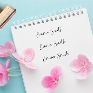 K&ouml;nnte beinhalten: Ein wei&szlig;es Notizbuch mit Spiralbindung und drei Zeilen Text, die in Schreibschrift "Emma Smith" lauten. Das Notizbuch liegt auf einer hellblauen Oberfl&auml;che mit rosa Bl&uuml;tenbl&auml;ttern.