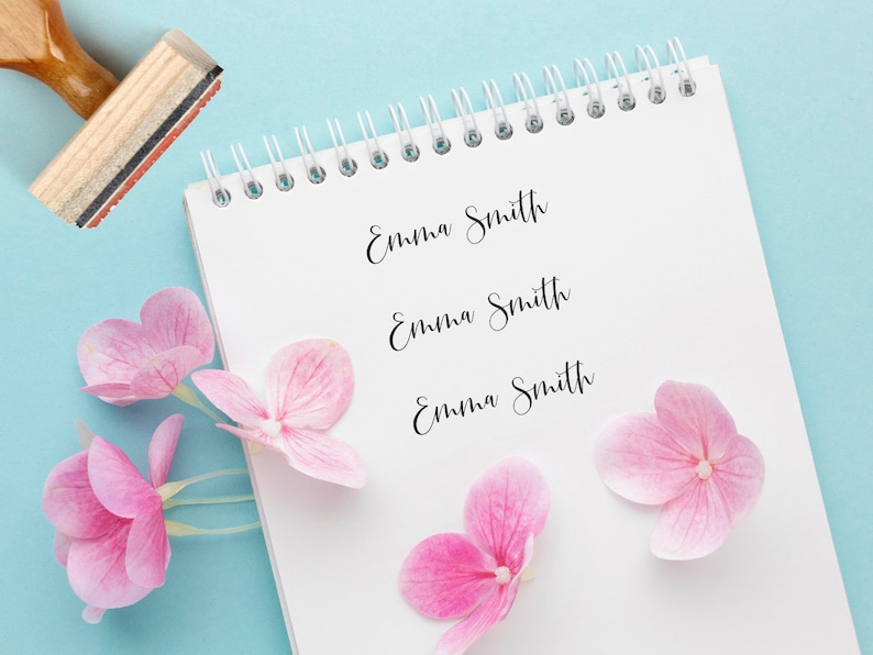 K&ouml;nnte beinhalten: Ein wei&szlig;es Notizbuch mit Spiralbindung und drei Zeilen Text, die in Schreibschrift "Emma Smith" lauten. Das Notizbuch liegt auf einer hellblauen Oberfl&auml;che mit rosa Bl&uuml;tenbl&auml;ttern.