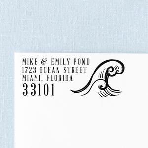 May include: A black ink stamp impression on white paper featuring the text MIKE & EMILY POND, 1723 OCEAN STREET, MIAMI, FLORIDA, 33101, accompanied by a stylized black graphic illustration of a crashing ocean wave to the right of the address.