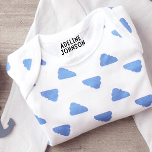 K&ouml;nnte beinhalten: Ein wei&szlig;er Babybody mit einem sich wiederholenden Muster aus hellblauen Wolken. Im Bereich des Halsausschnitts ist der Name ADELINE JOHNSON in einer fetten, schwarzen, serifenlosen Schrift aufgedruckt. Das Kleidungsst&uuml;ck hat kurze &Auml;rmel.