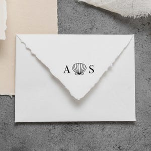 May include: A white envelope with a deckled edge flap featuring a black ink stamp of the letter A, a seashell icon, and the letter S centered on the point of the flap. The envelope is rectangular and made of thick, textured paper stock.