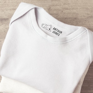 K&ouml;nnte beinhalten: Ein wei&szlig;er Babybody mit einem schwarzen Grafikdruck am inneren Halsausschnitt. Der Druck besteht aus einer kleinen Illustration eines schlafenden Welpen links neben dem Text ARTHUR JONES, der in einer fetten serifenlosen Schrift gedruckt ist.