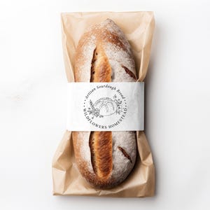 May include: An oval loaf of sourdough bread with a golden-brown crust and a central score, wrapped in brown paper and secured with a white paper band featuring the text Artisan Sourdough Bread and WILDFLOWERS HOMESTEAD with a floral illustration.