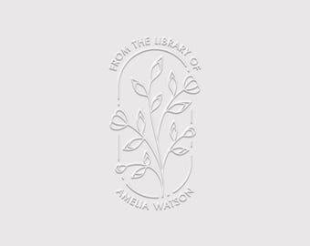May include: A white embossed stamp design featuring a central botanical illustration of a leafy branch with small flowers. The design is enclosed in an oval frame with the text FROM THE LIBRARY OF arched at the top and AMELIA WATSON at the bottom.
