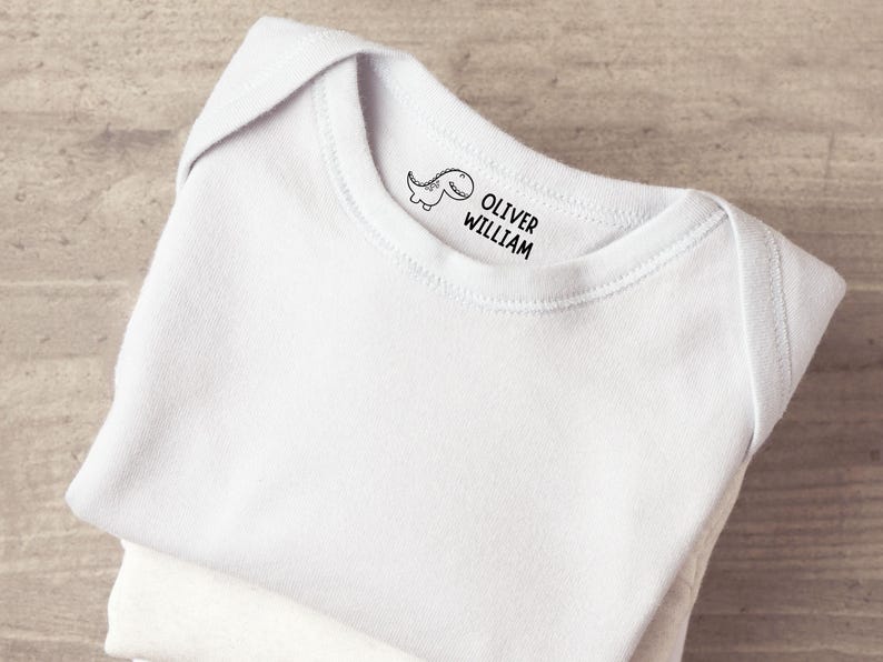 K&ouml;nnte beinhalten: Ein wei&szlig;er Babybody mit einem schwarzen Stempelaufdruck am inneren Halsausschnitt. Der Aufdruck zeigt eine kleine Cartoon-Dinosaurier-Grafik neben dem Text OLIVER WILLIAM in einer einfachen, fetten, serifenlosen Schrift zur Kennzeichnung.