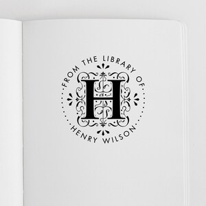 May include: A black ink stamp impression on white paper featuring a central letter H surrounded by ornate scrollwork. The circular design includes the text FROM THE LIBRARY OF and HENRY WILSON, framed by a dotted border with small decorative accents.