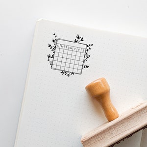 May include: A wooden stamp with a black ink pad and a design of a calendar with the days of the week labeled "SMTWTFS" surrounded by floral vines.