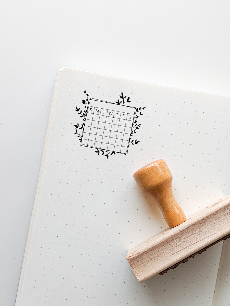 May include: A wooden stamp with a black ink pad and a design of a calendar with the days of the week labeled "SMTWTFS" surrounded by floral vines.