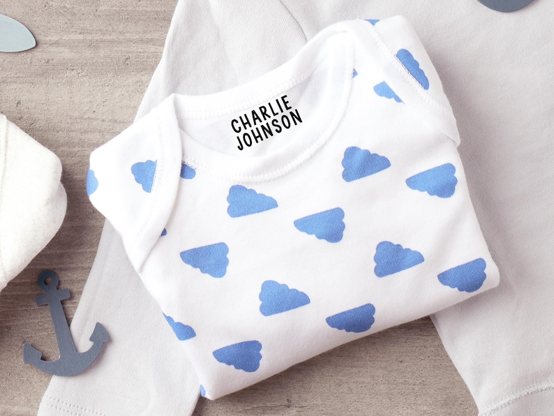 K&ouml;nnte beinhalten: Ein wei&szlig;er, kurz&auml;rmeliger Babybody mit einem sich wiederholenden Muster aus hellblauen Wolken. Im Bereich des Halsausschnitts ist der Name CHARLIE JOHNSON in fetten, schwarzen Gro&szlig;buchstaben aufgedruckt. Das Kleidungsst&uuml;ck hat einen Rundhalsausschnitt.