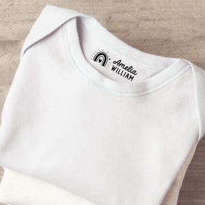 K&ouml;nnte beinhalten: Ein wei&szlig;er Babybody aus Baumwolle mit einem schwarzen Aufdruck am inneren Halsausschnitt. Der Druck zeigt ein kleines Regenbogensymbol mit einem Herz in der Mitte, gefolgt von dem Text Amelia WILLIAM in einer Kombination aus Schreib- und Blockschrift.