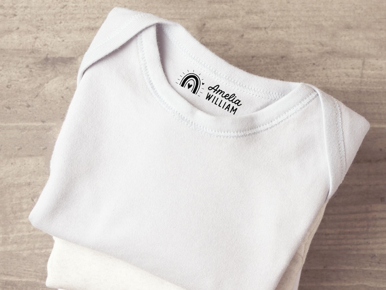 K&ouml;nnte beinhalten: Ein wei&szlig;er Babybody aus Baumwolle mit einem schwarzen Aufdruck am inneren Halsausschnitt. Der Druck zeigt ein kleines Regenbogensymbol mit einem Herz in der Mitte, gefolgt von dem Text Amelia WILLIAM in einer Kombination aus Schreib- und Blockschrift.
