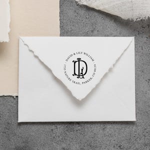May include: A white envelope with a deckled edge flap featuring a black circular ink stamp. The stamp displays the monogram DL in the center, surrounded by the text David & Lily William, 1755 Nature Trail, Parker, CO 80134 in a serif font.