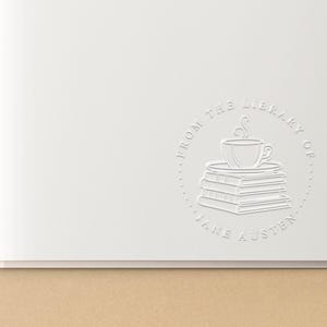 May include: A white embossed circular stamp on a page of an open book. The design features a stack of three books topped with a steaming cup of coffee, surrounded by the text FROM THE LIBRARY OF JANE AUSTEN in a circular border.