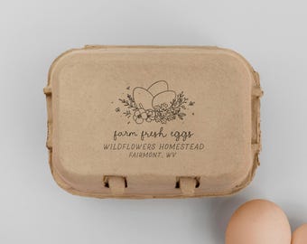 May include: A rectangular tan cardboard egg carton featuring a black ink stamp of three eggs nestled among wildflowers. The text reads farm fresh eggs, WILDFLOWERS HOMESTEAD, and FAIRMONT, WV. Two light brown eggs are visible in the lower right corner.