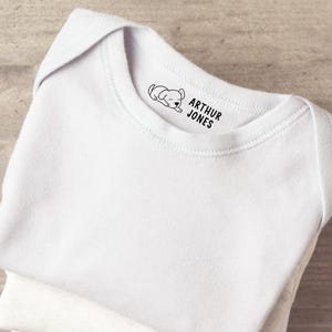 K&ouml;nnte beinhalten: Ein wei&szlig;er Babybody mit einem schwarzen Grafikdruck am inneren Halsausschnitt. Der Druck besteht aus einer kleinen Illustration eines schlafenden Welpen links neben dem Text ARTHUR JONES, der in einer fetten serifenlosen Schrift gedruckt ist.