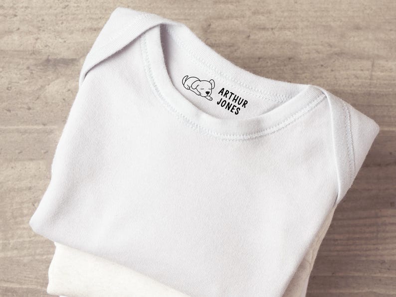 K&ouml;nnte beinhalten: Ein wei&szlig;er Babybody mit einem schwarzen Grafikdruck am inneren Halsausschnitt. Der Druck besteht aus einer kleinen Illustration eines schlafenden Welpen links neben dem Text ARTHUR JONES, der in einer fetten serifenlosen Schrift gedruckt ist.